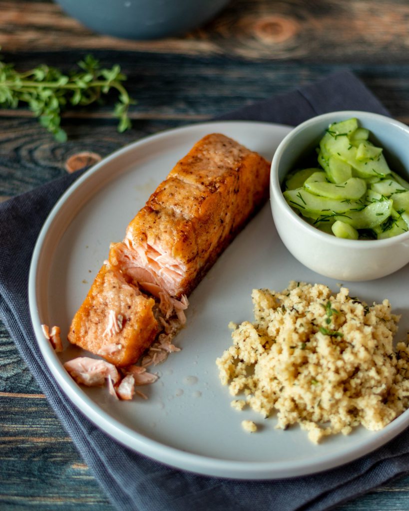 Gebratenes Lachsfilet mit Couscous auf einem Teller. Dazu eine Schüssel Gurkensalat