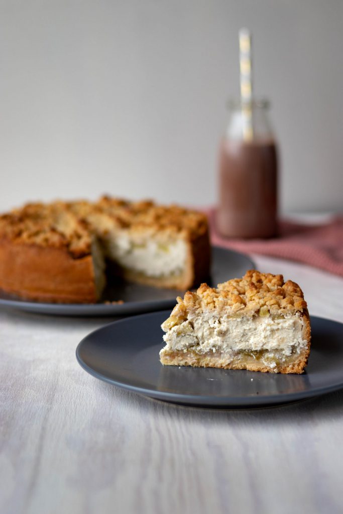 Ein Stück Rhabarberkuchen im Vordergrund. Im Hintergrund ein ganzer Kuchen.