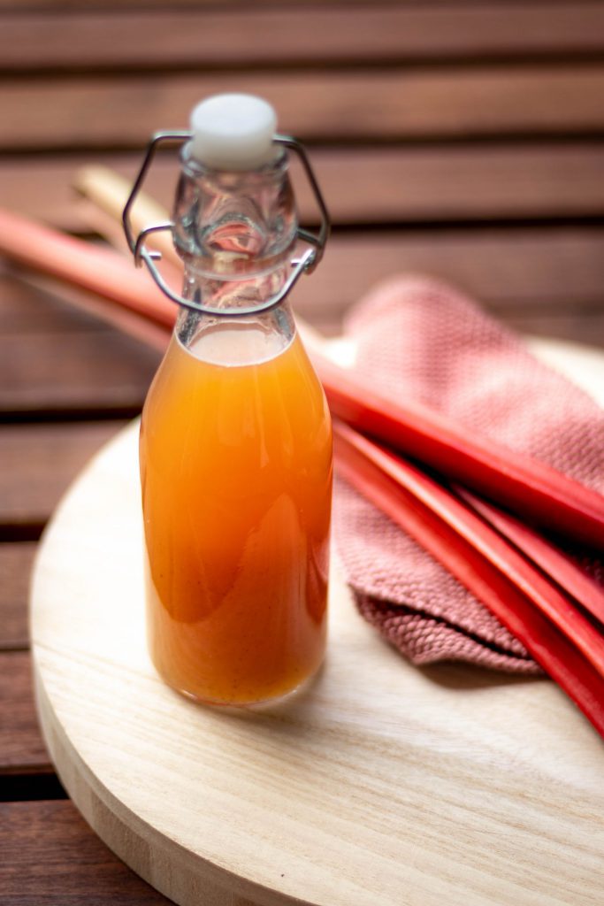 Eine Flasche Rhabarbersirup auf einem Holztablett. Daneben liegen drei Stangen Rhabarber