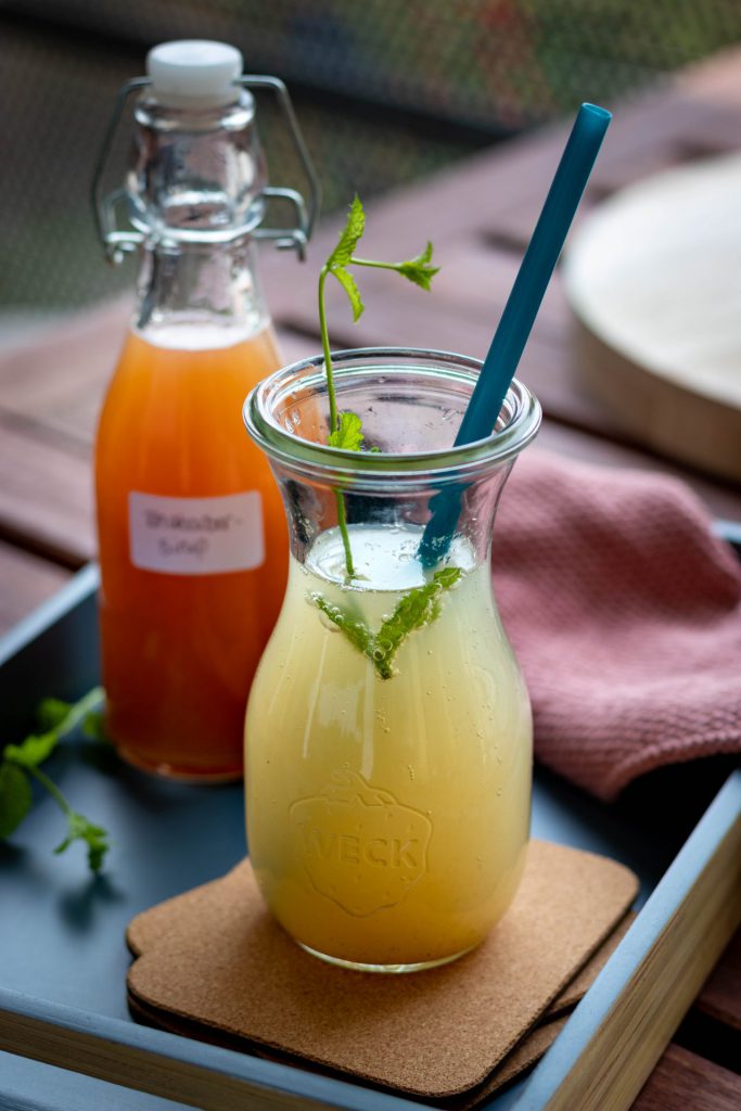 Ein Glas Rhabarberlimonade mit einem Stängel Minze und einem Strohhalm. Im Hintergrund eine Flasche Rhabarbersirup