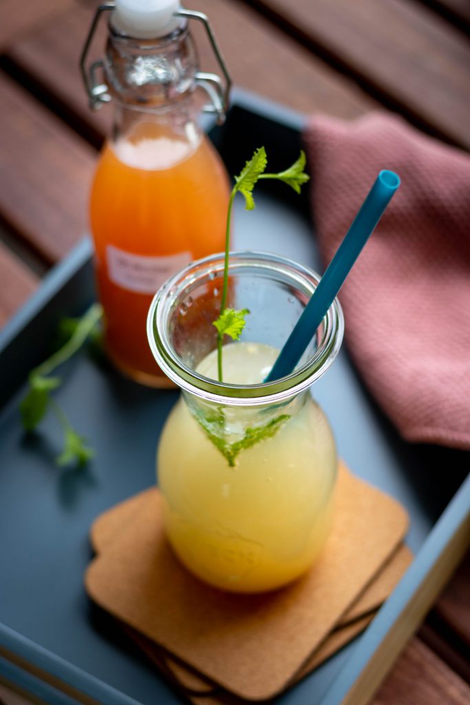 Ein Glas Rhabarberlimonade mit einem Stängel Minze und einem Strohhalm. Im Hintergrund eine Flasche Rhabarbersirup